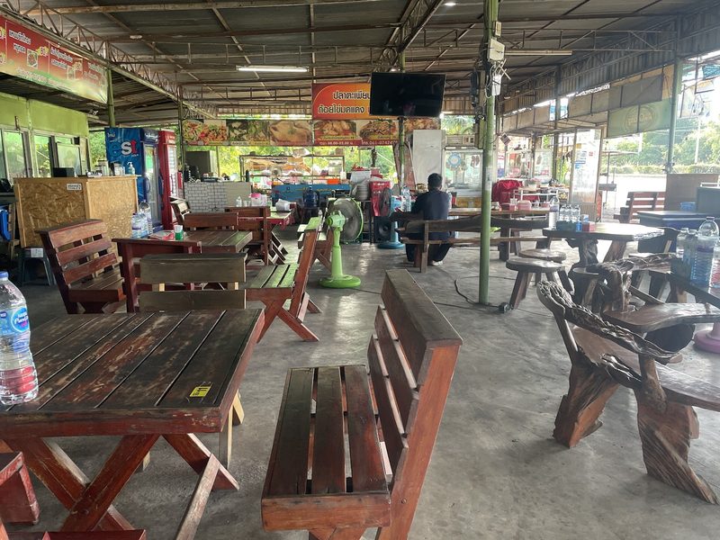 ในร้านลูกชายกำนันบุญส่ง โต๊ะและม้านั่งไม้ใต้หลังคาโปร่ง ป้ายเมนูเหนือครัวด้านหลัง
